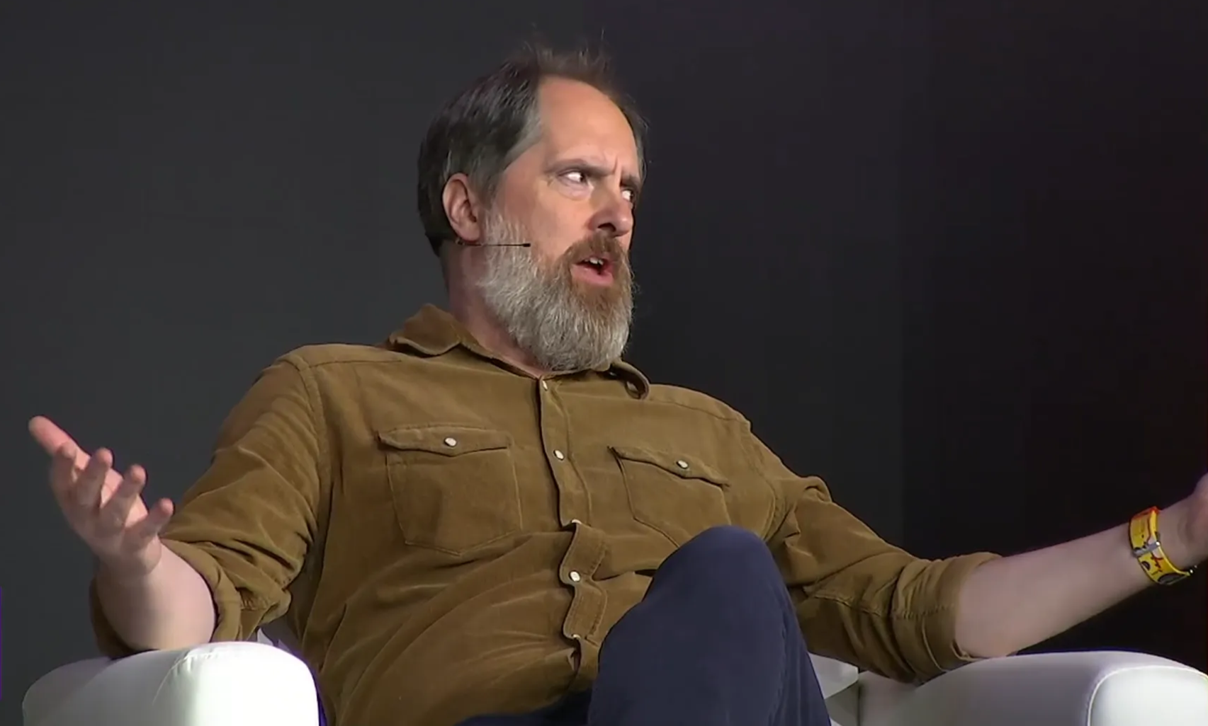 Bearded actor Brendan Hunt puts his arms out wide while sitting and talking to the audience